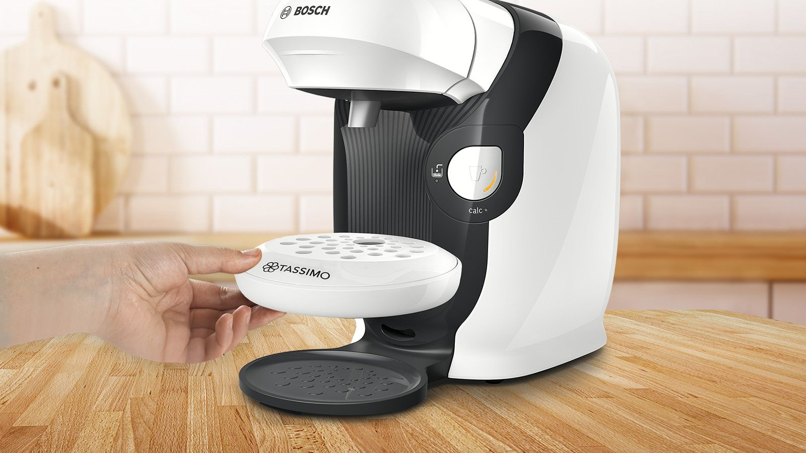 Eine weiße Tassimo Kaffeemaschine. Eine Hand hält einen weißen Tassimo-Disc-Halter. Holztisch.