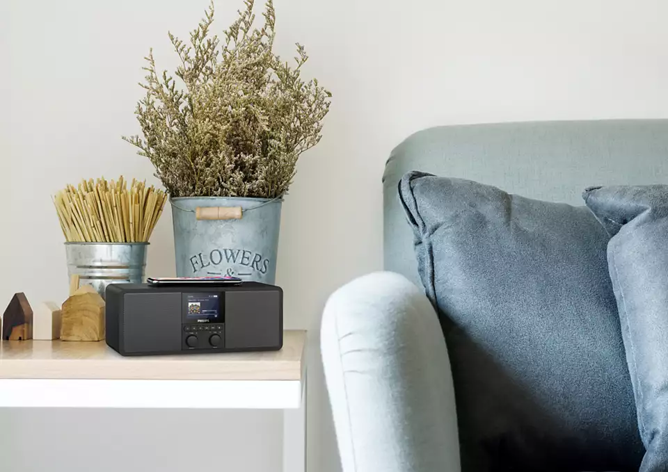 Radio op een tafel met bloemen, een telefoon en een bank op de achtergrond.