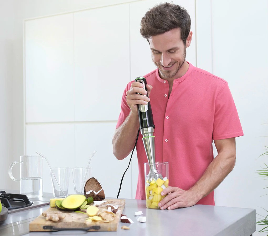 Mann mixt Mango in einem Glas mit einem Stabmixer auf einer Theke, Zutaten sind drumherum.