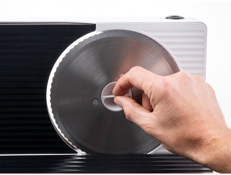A hand adjusts the white plastic part of a meat slicer. The blade is stainless steel, reflecting light. The slicer is white and black.