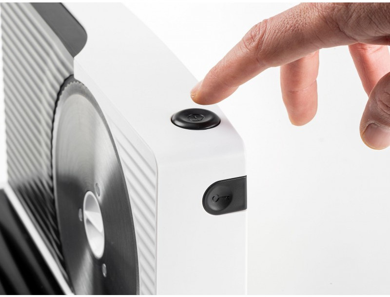 A hand presses a black button on a white food slicer. A circular blade is visible, and the background is white.