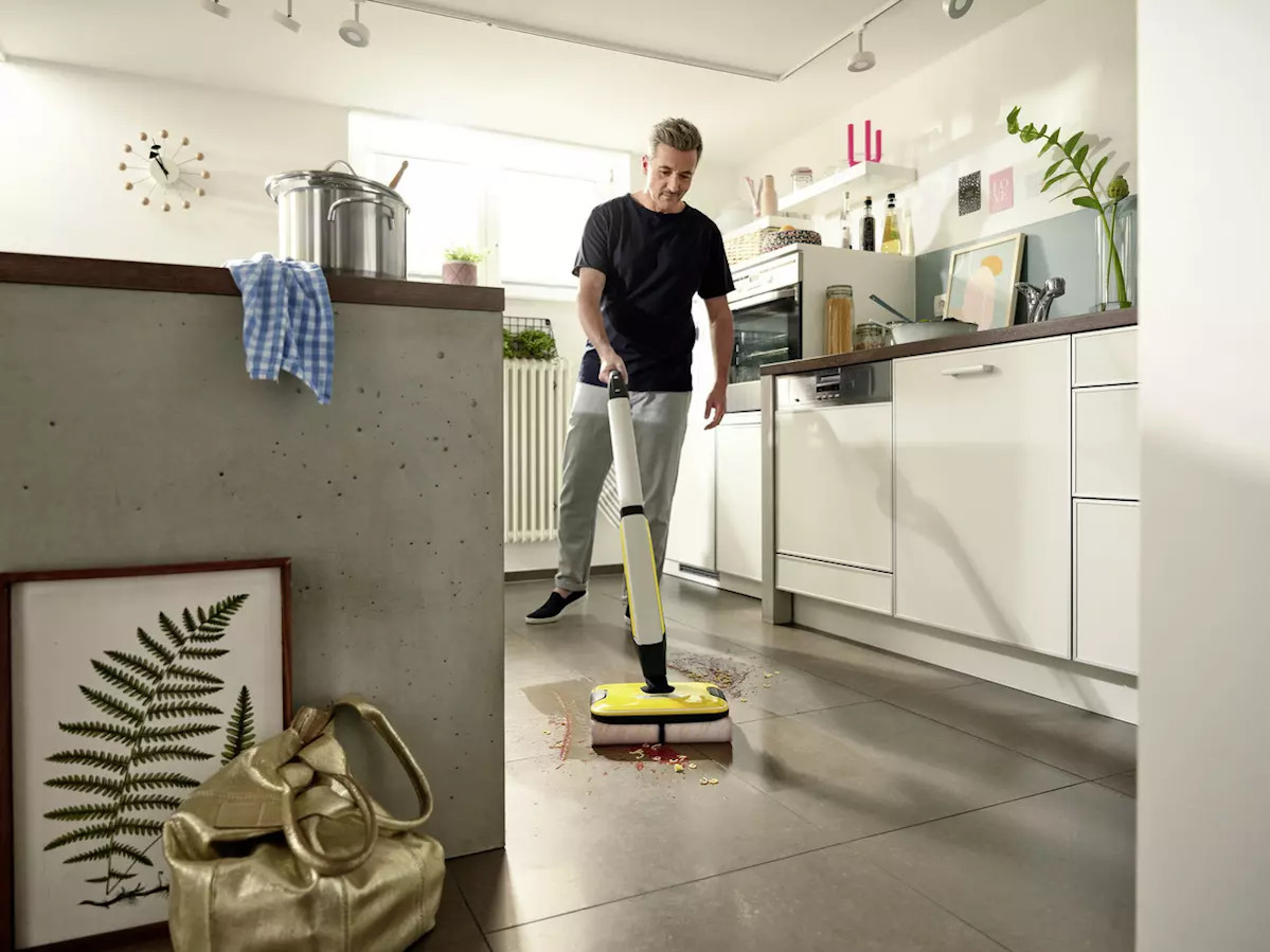 Un homme nettoie le sol de la cuisine avec une machine de nettoyage de sol jaune et noire. La scène comprend un comptoir et des armoires.