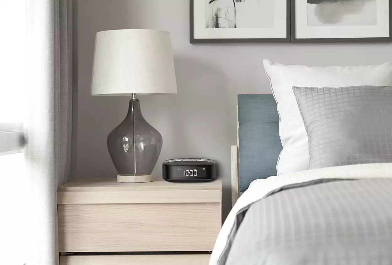 Bedroom scene with a lamp, alarm clock, and bed with pillows and a blanket.