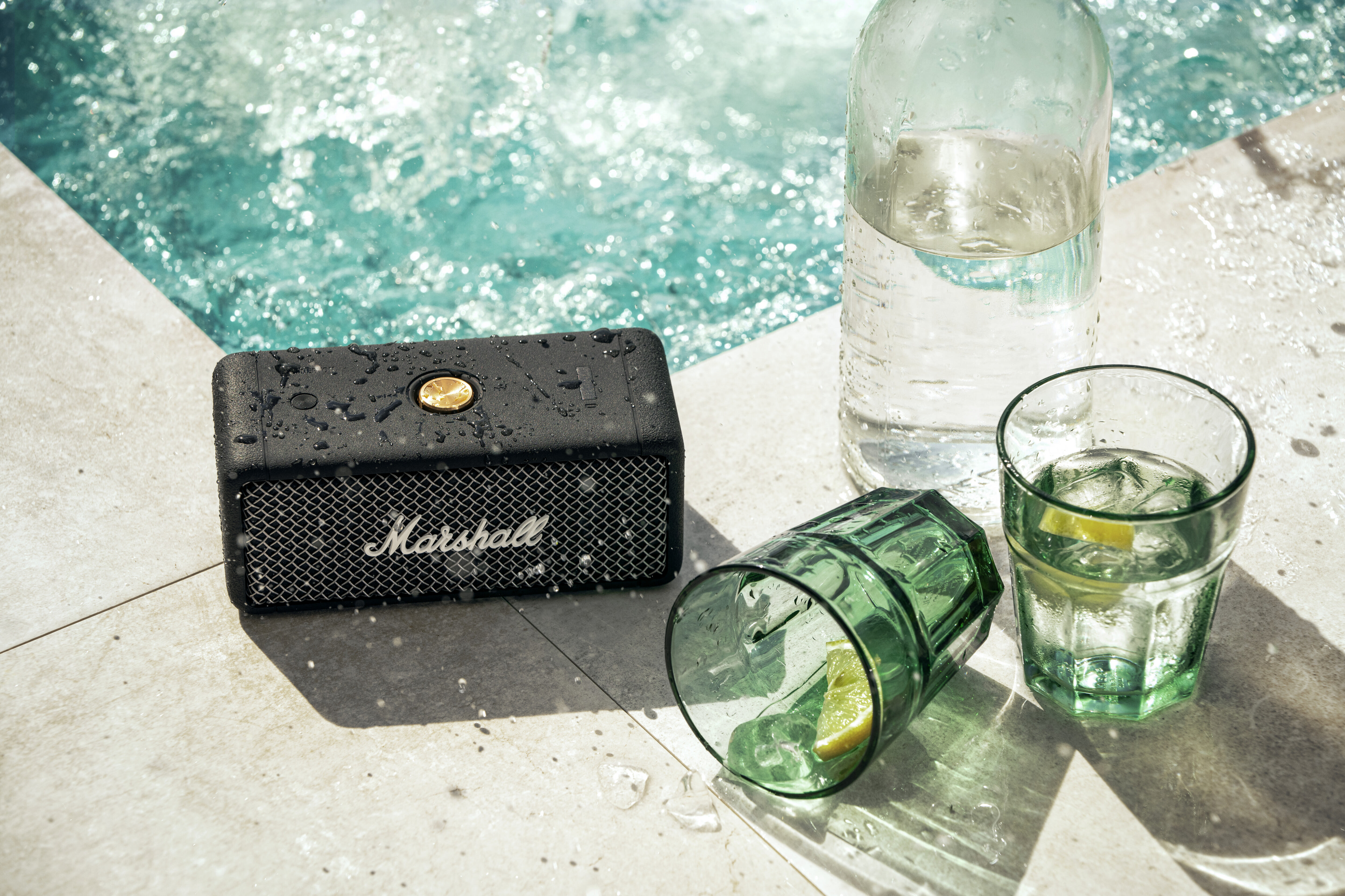Ein schwarzer Marshall-Lautsprecher, eine Wasserflasche und zwei grüne Gläser mit Eis und Zitrone auf einem Pooldeck.