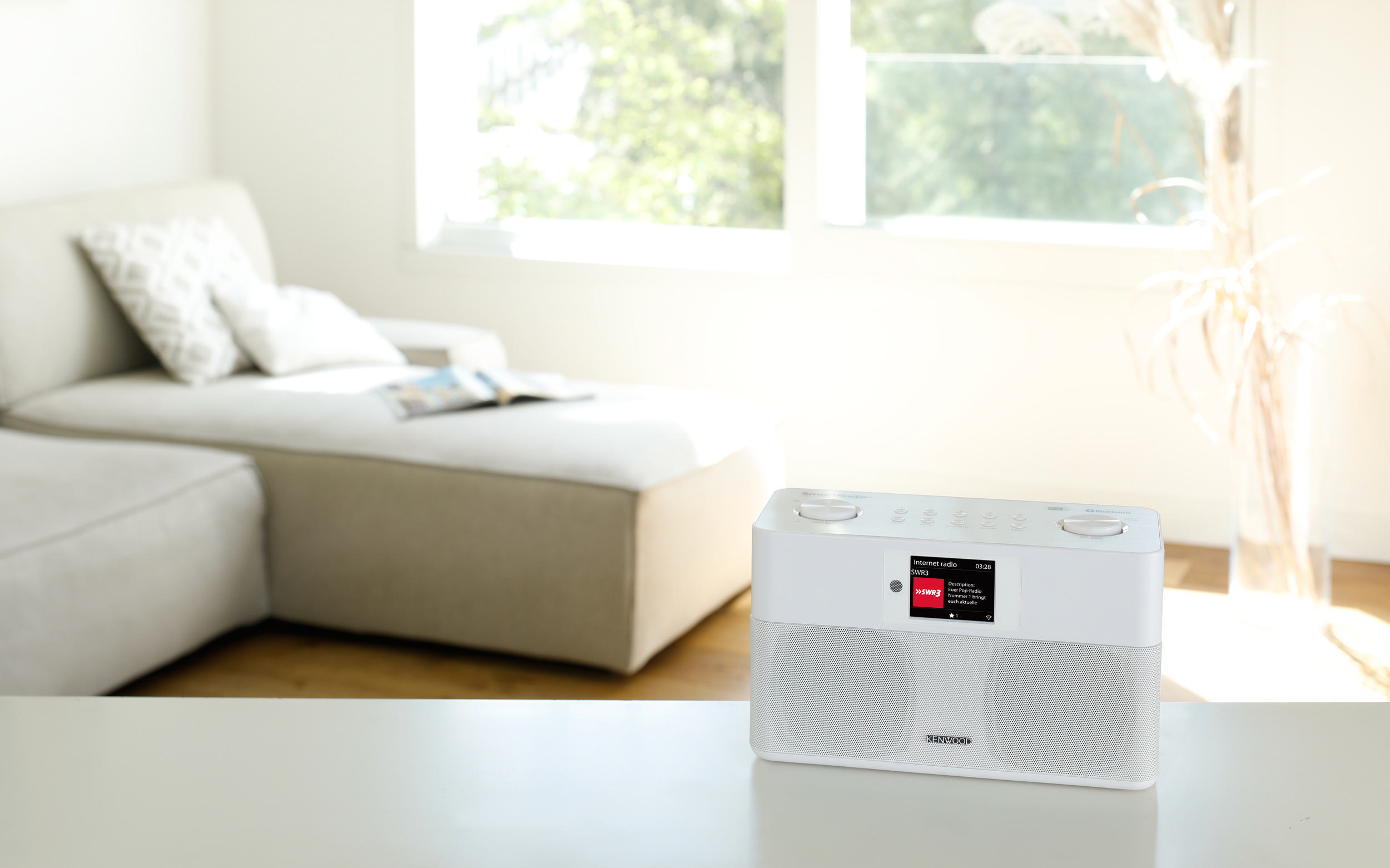 Weißes Radio auf einem Tisch. Eine Couch und ein Fenster befinden sich im Hintergrund und bilden ein Wohnzimmer.
