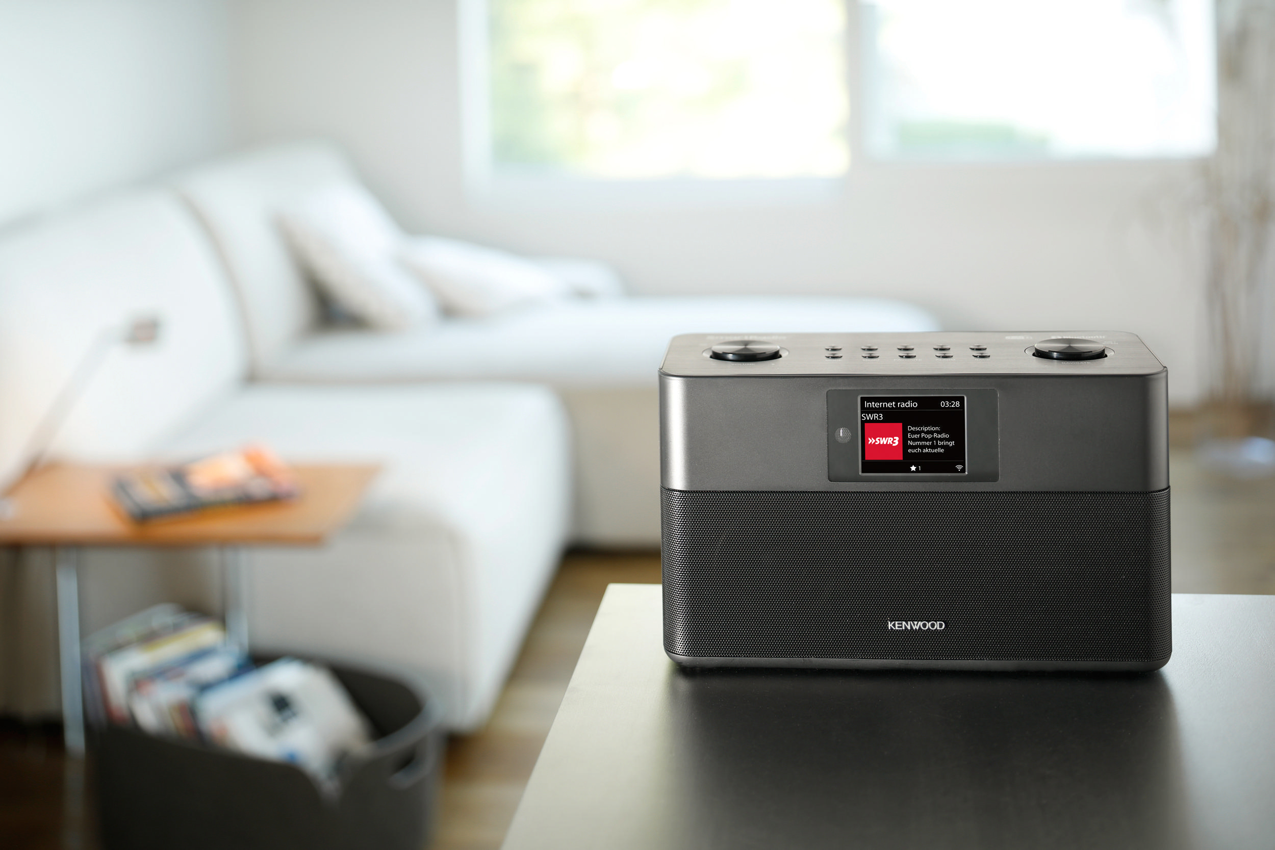 Ein Kenwood Internetradio auf einem Tisch, das einen Bildschirm mit Radiosenderinformationen anzeigt. Ein Sofa im Hintergrund.