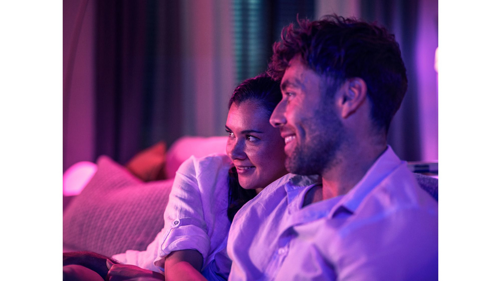 Pareja sonriendo, iluminada con luz púrpura, sentada en un sofá.