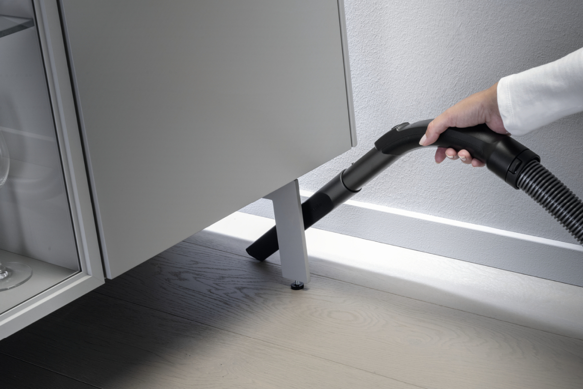 Eine Person, die mit einem schwarzen Schlauch unter einem Schrank saugt.