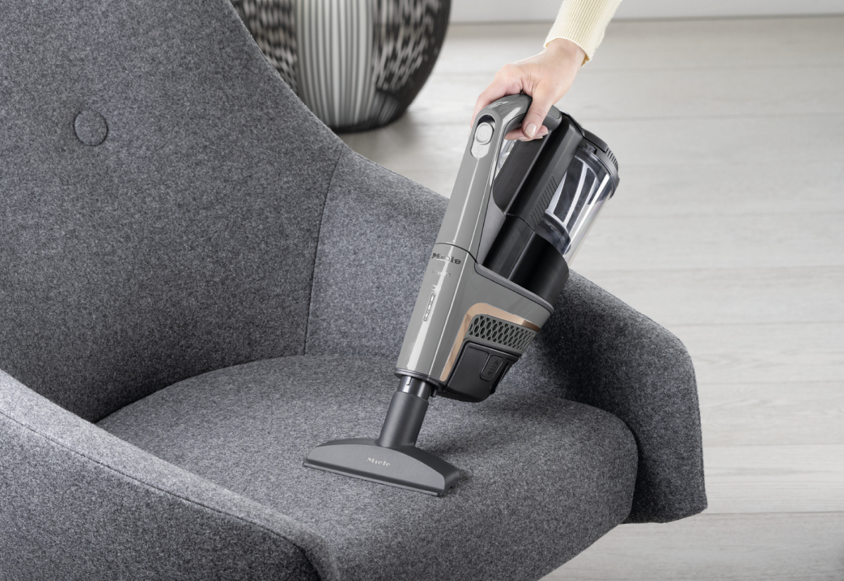 Eine Person saugt einen grauen Stuhl mit einem grau-schwarzen Handstaubsauger ab.