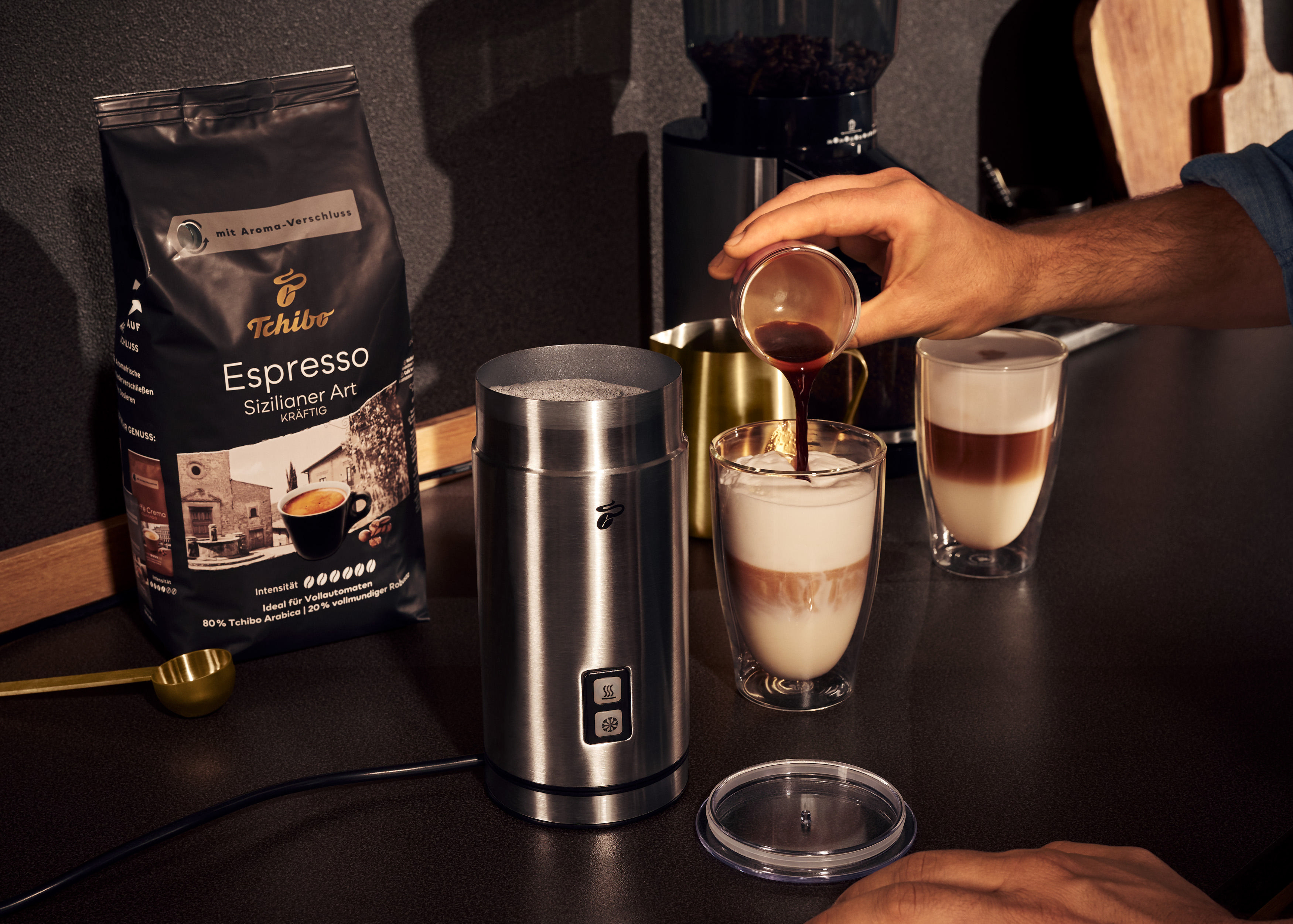 Eine Person gießt Kaffee in ein Glas. Kaffeemaschine und Kaffeebohnen stehen auf dem Tisch.