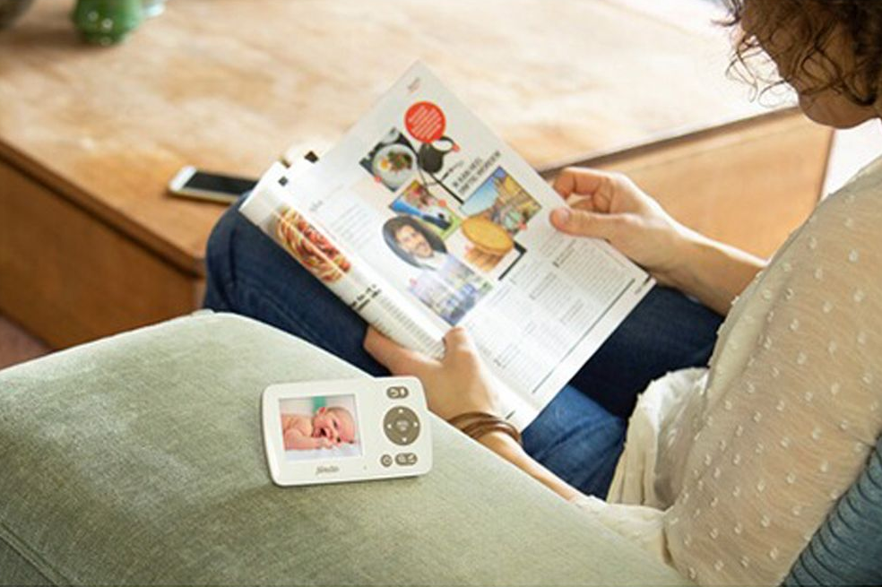 Femme lisant un magazine tout en regardant un babyphone montrant un bébé dans un berceau.