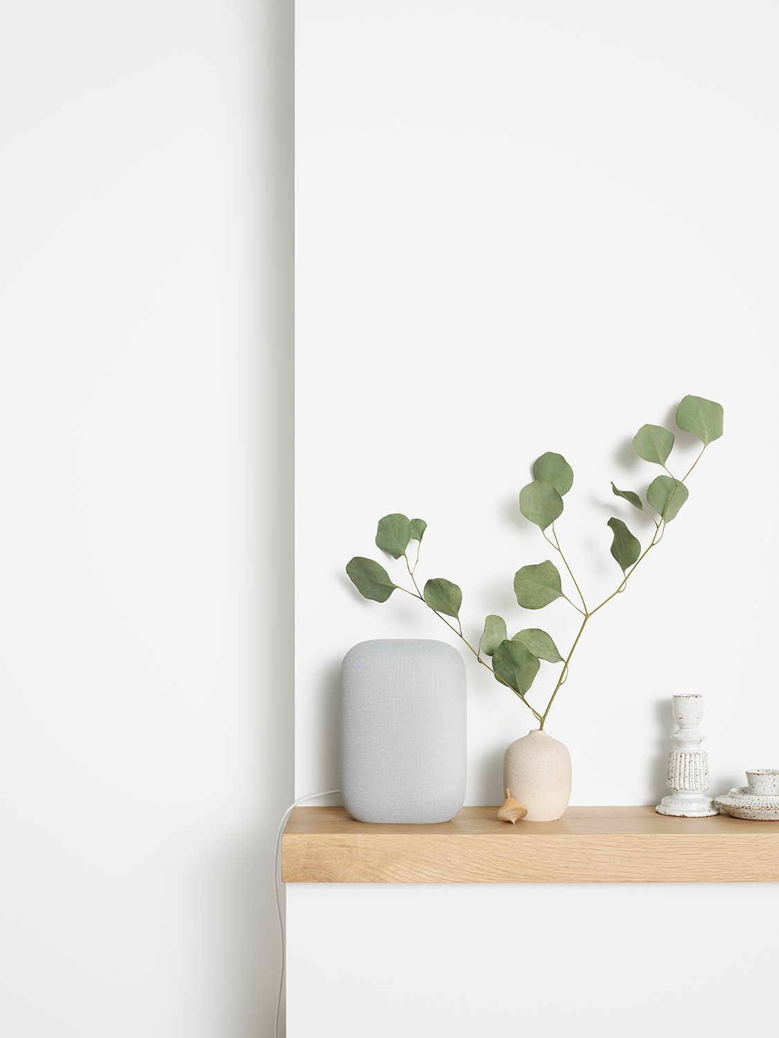 Een grijze luidspreker, vaas met groene bladeren en kandelaar op een houten plank tegen een witte muur.