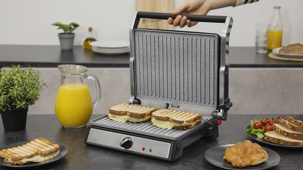 Una plancha para sándwiches con dos sándwiches dentro. Hay zumo de naranja y otros alimentos.