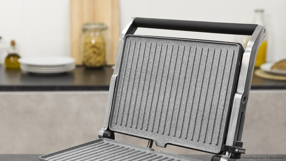 Parrilla eléctrica abierta con placas de parrilla grises. Ambiente de cocina con platos, aceite y pasta al fondo.