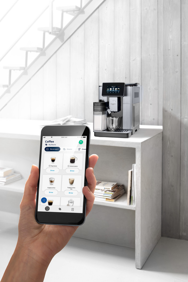 Hand, die ein Telefon mit Kaffeemaschinen-App hält, daneben eine Kaffeemaschine.