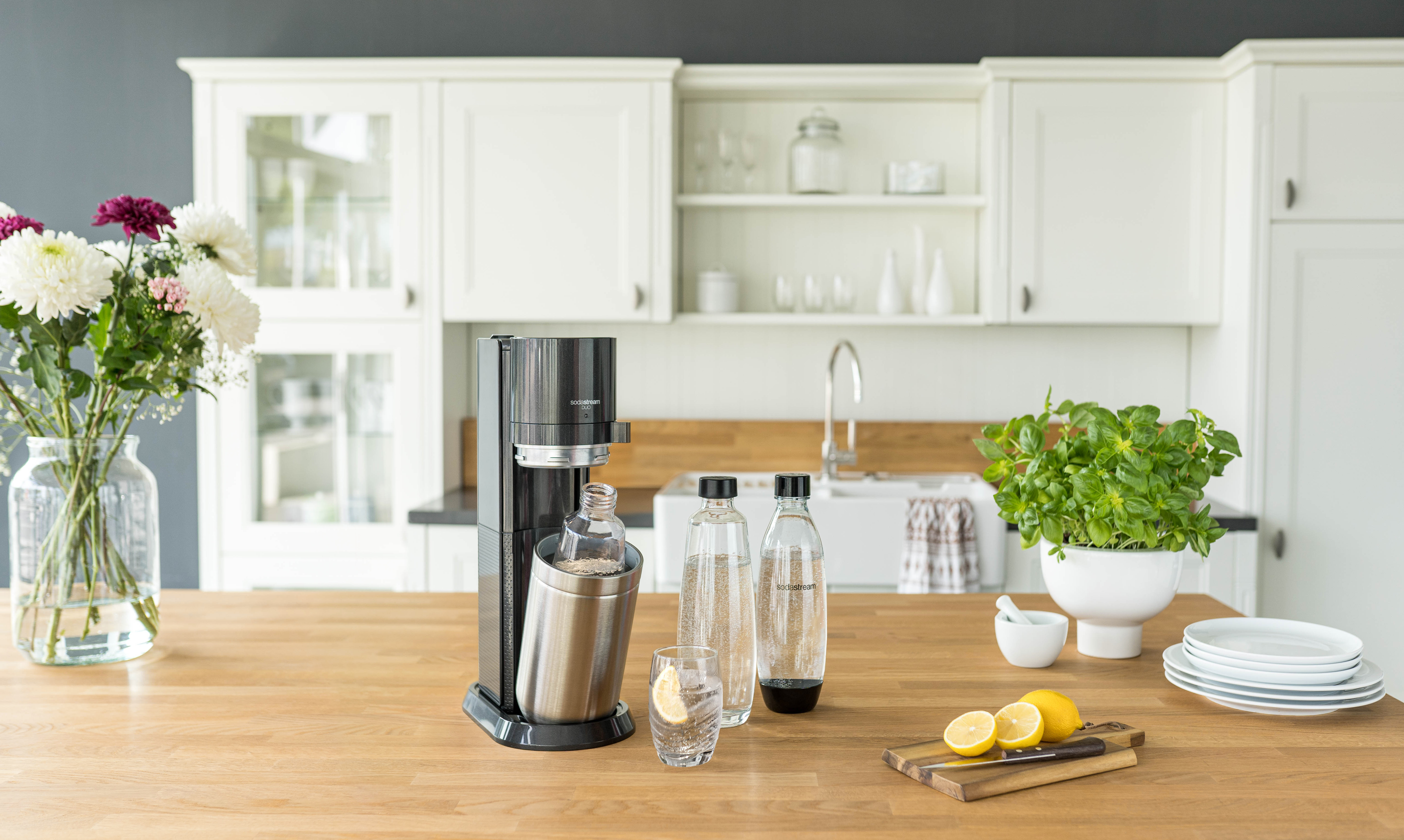 Küche mit Soda-Maker, Wasserflaschen, Blumen und Zitronen auf Holztheke.