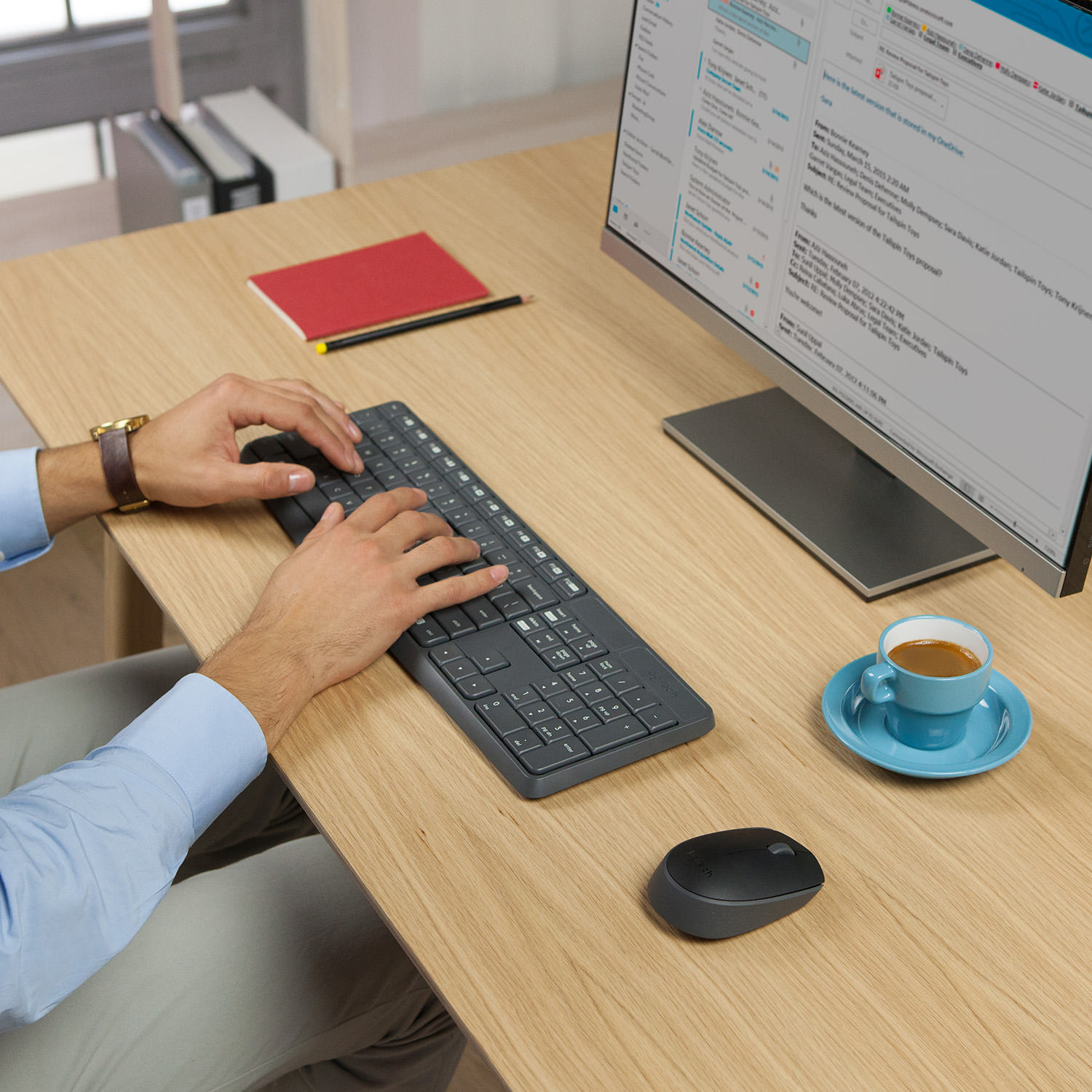 Person verwendet Tastatur, Maus und Monitor auf einem Holztisch mit Kaffeetasse.