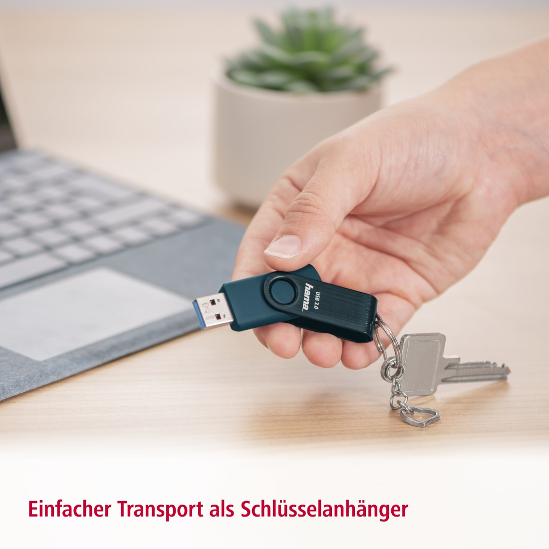 Hand hält USB-Stick mit Schlüsseln auf Holzschreibtisch. Laptop und Pflanze im Hintergrund.