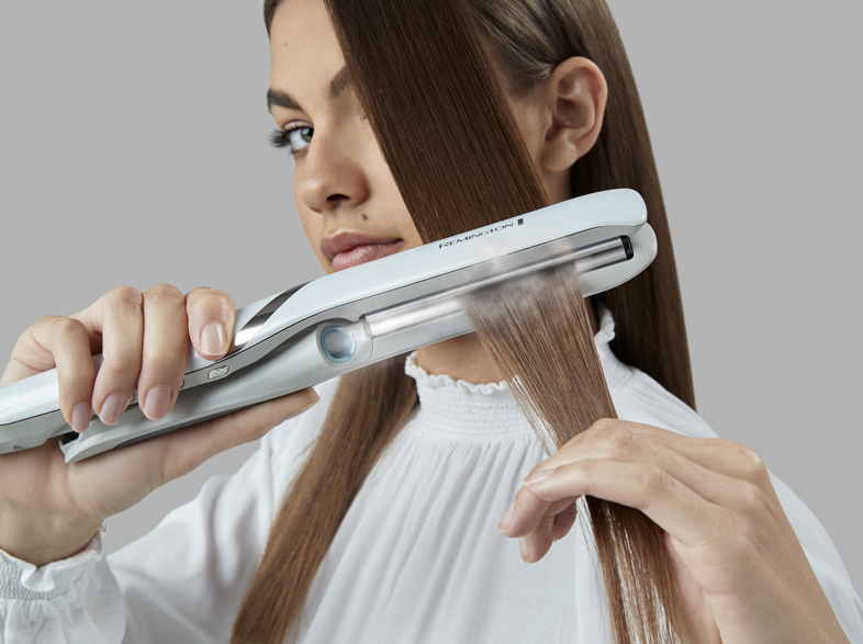 Egy nő fehér Remington hajvasalóval egyenesíti a haját. A haj barna és hosszú.