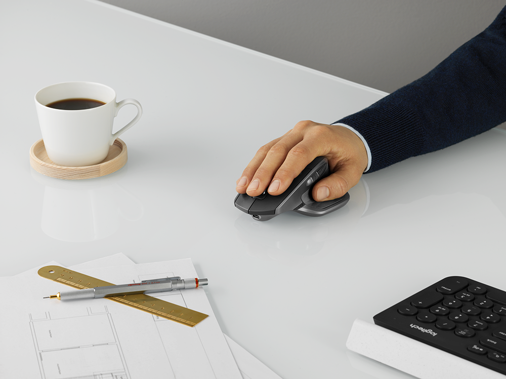 Une personne utilise une souris grise sur un bureau blanc, avec une tasse de café et des fournitures de bureau.