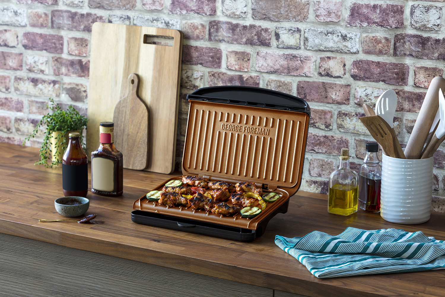 George Foreman grill with cooked food. Ingredients and utensils around, set against a brick wall background.
