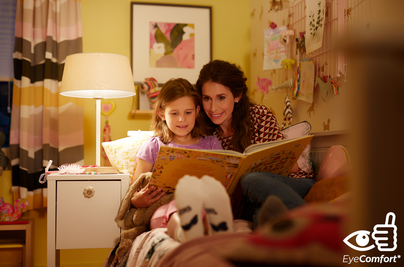 Eine Frau und ein Mädchen lesen gemeinsam ein Buch in einem Schlafzimmer, beleuchtet von einer Lampe.