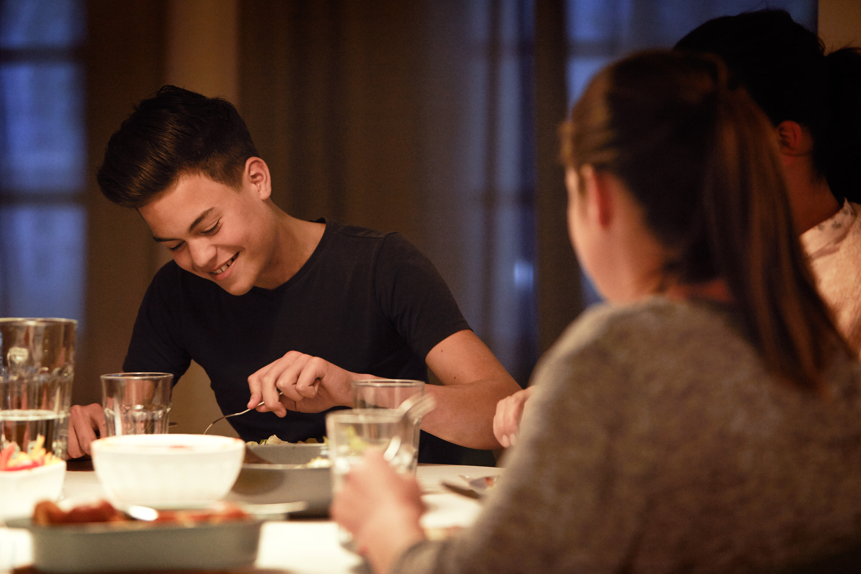Teenager beim Abendessen lächelt, isst. Andere am Tisch, Teller, Gläser, dunkler Raum.