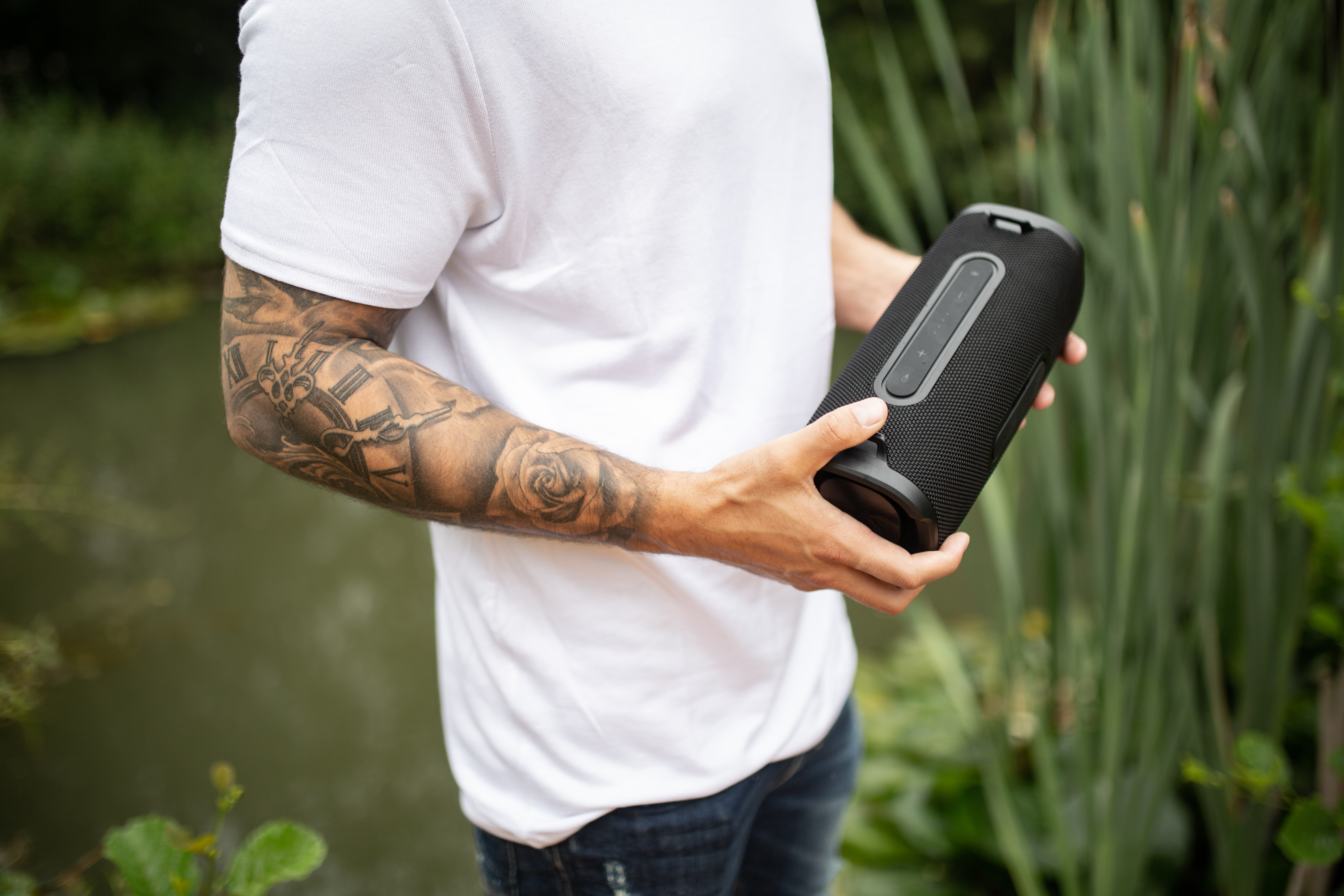 Ein Mann hält einen schwarzen Lautsprecher mit einem Tattoo auf dem Arm, steht in der Nähe von Wasser und Grün.