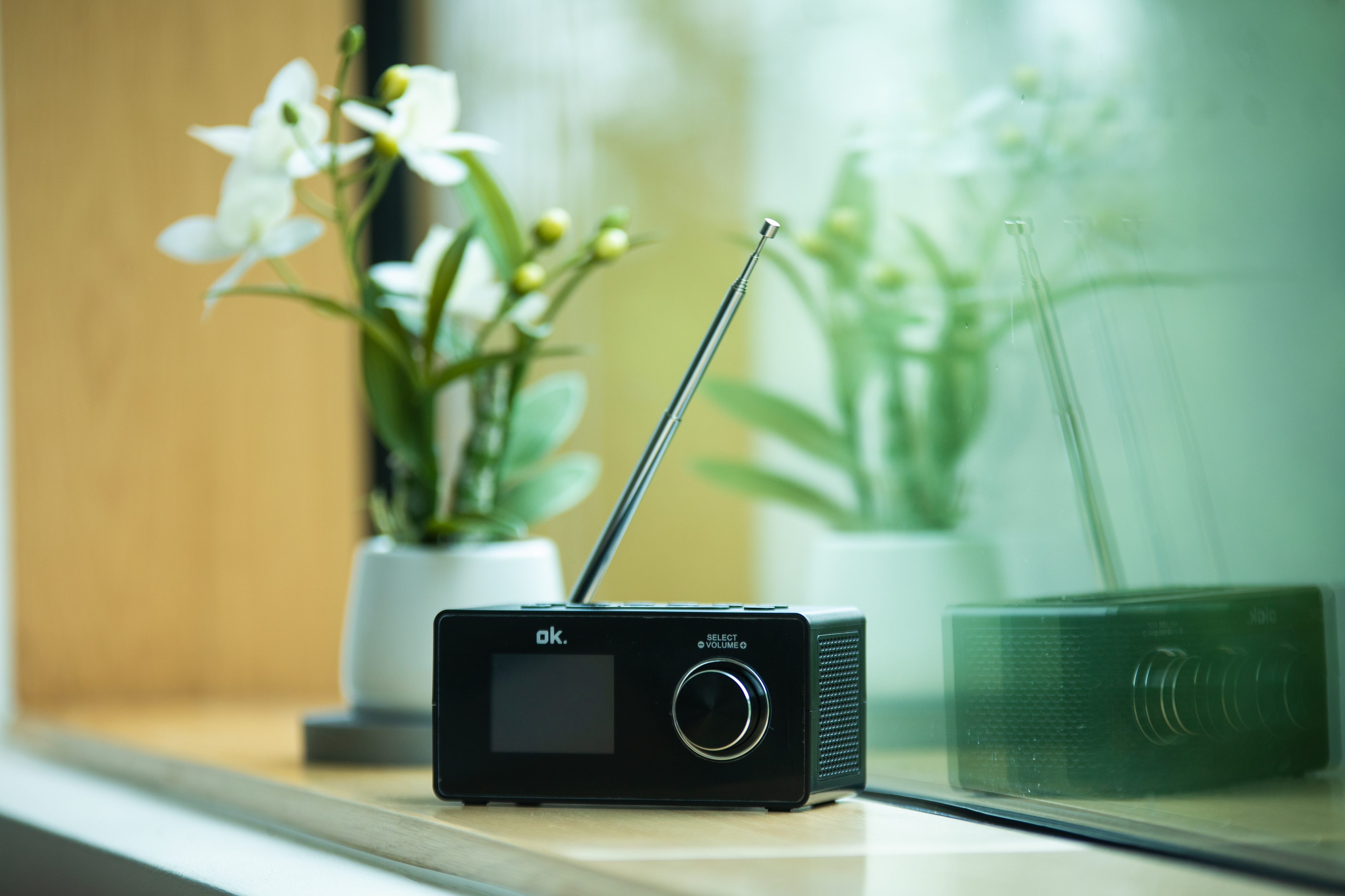Schwarzes Radio mit Antenne auf Fensterbank, weiße Blumen im Hintergrund. Fensterspiegelung.