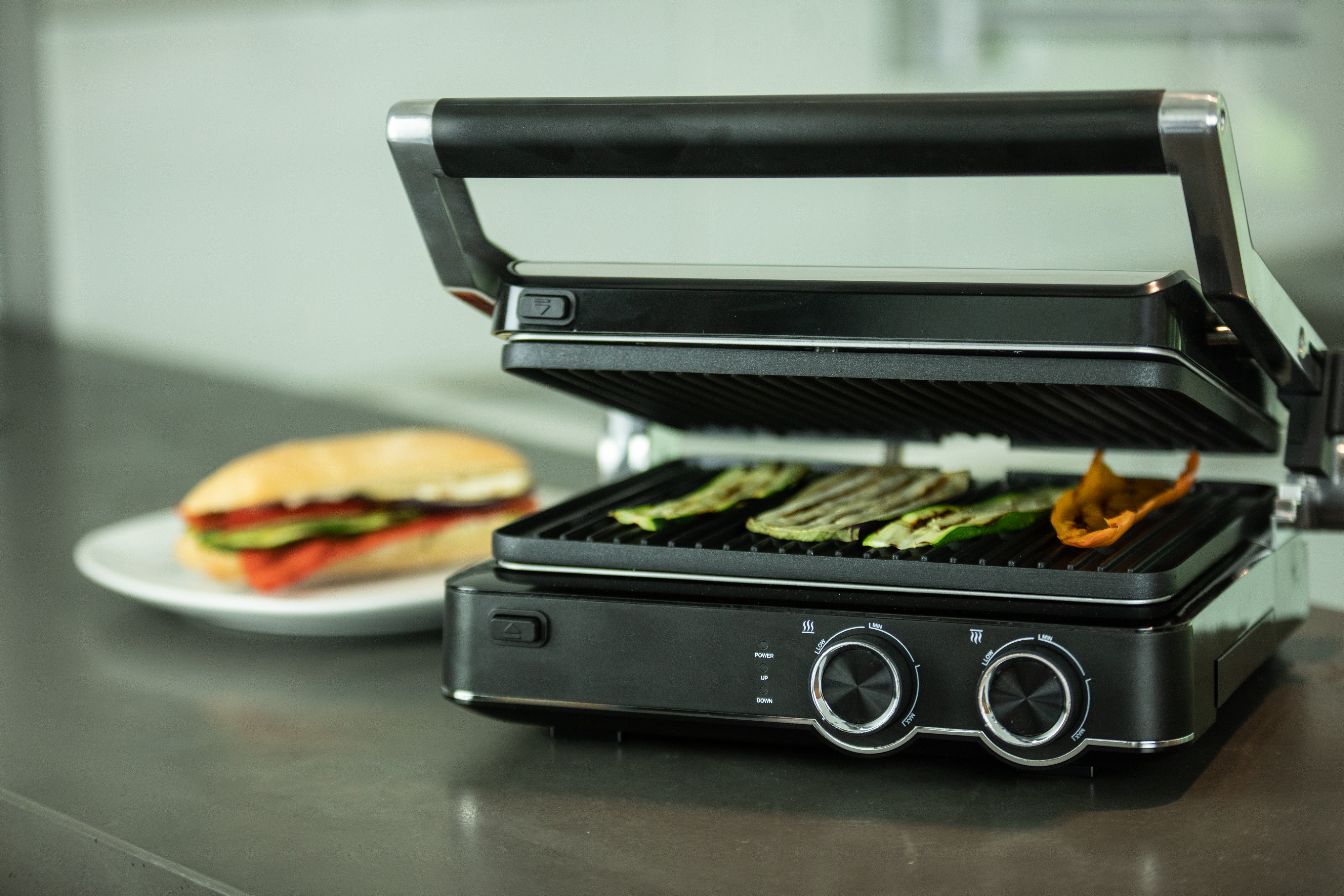 Ein schwarzer Grill mit Essen auf dem Grill und einem Sandwich auf einem Teller links.