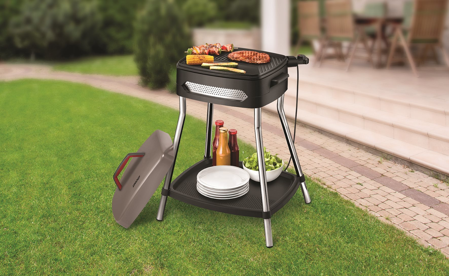 Fekete grill állványon, rajta étellel, zöld gyep és kőút mellett. A fedél nyitva.