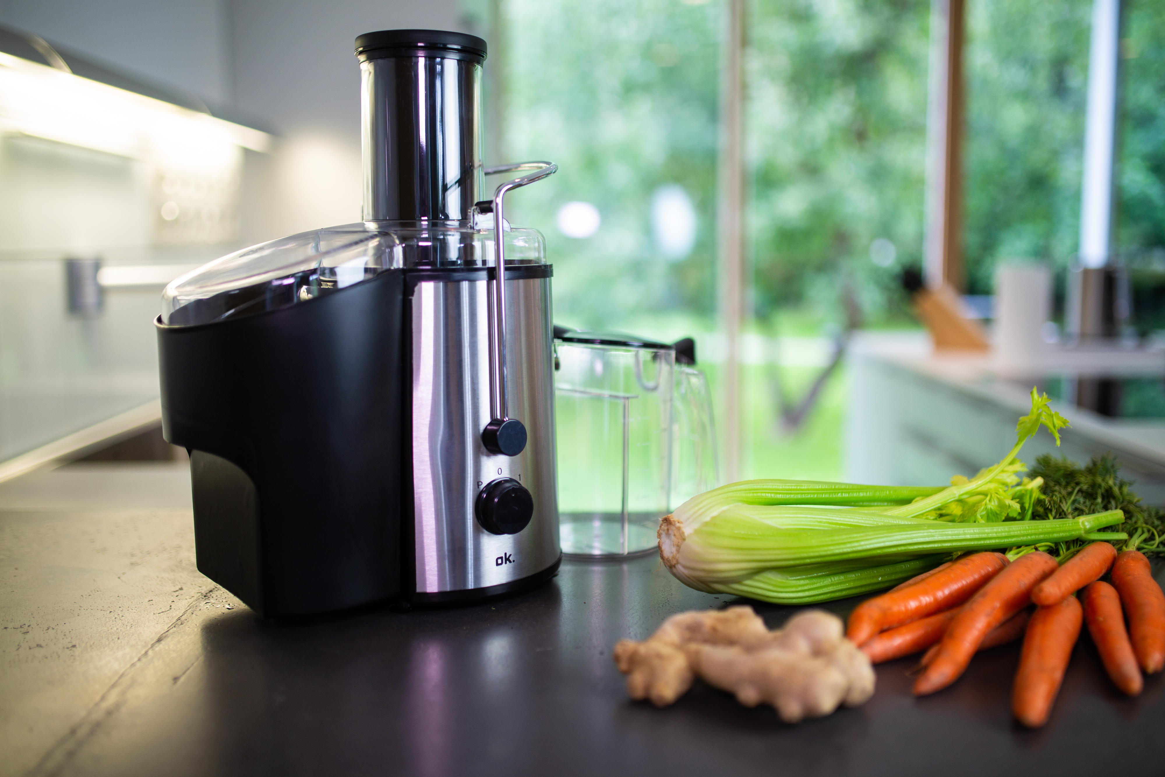 Entsafter mit Karotten, Sellerie, Ingwer auf schwarzer Theke, Fenster im Hintergrund.
