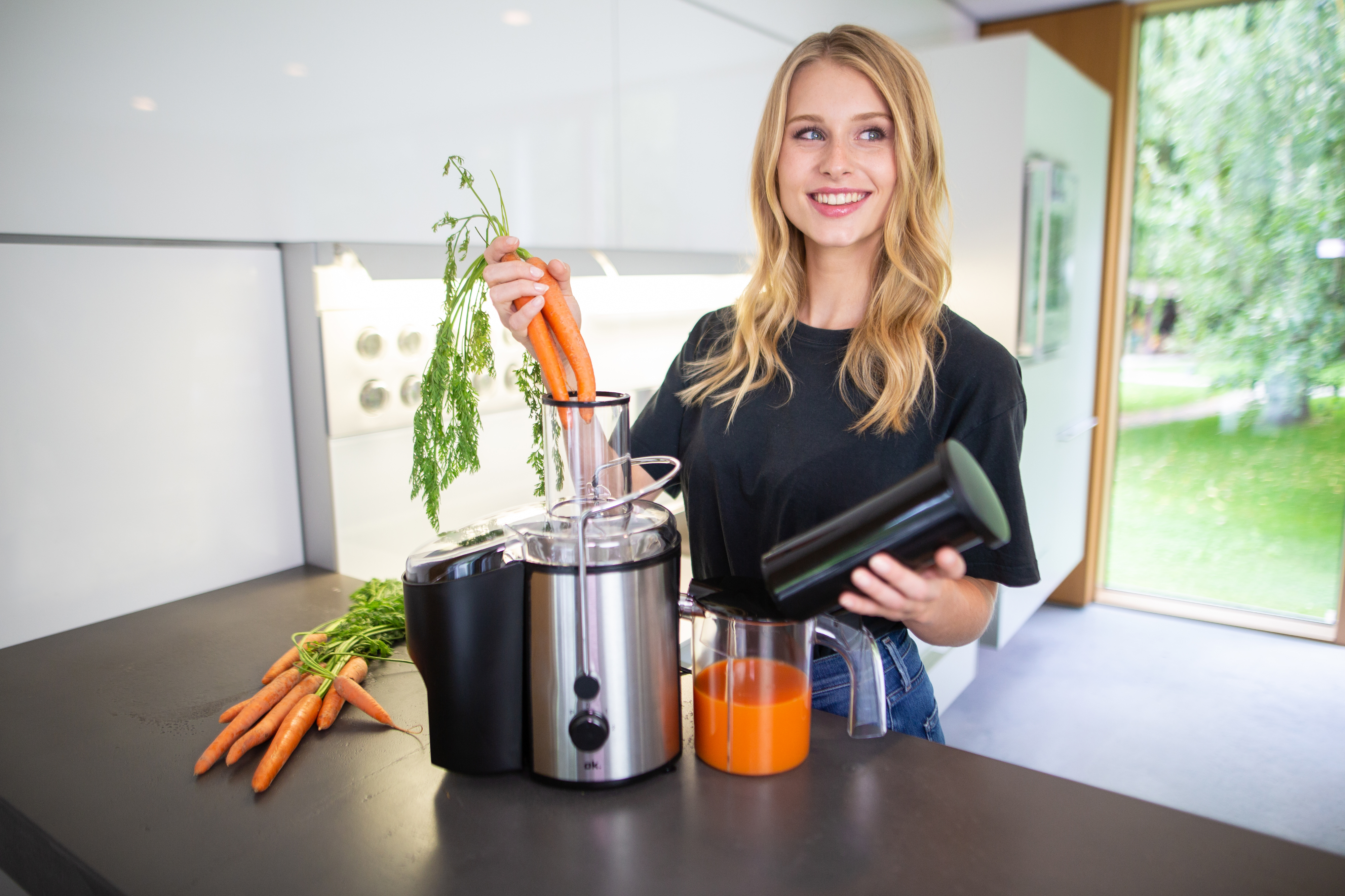 Eine Frau lächelt, hält Karotten an einem Entsafter. Karotten sind auf der Theke, Saft im Glas.