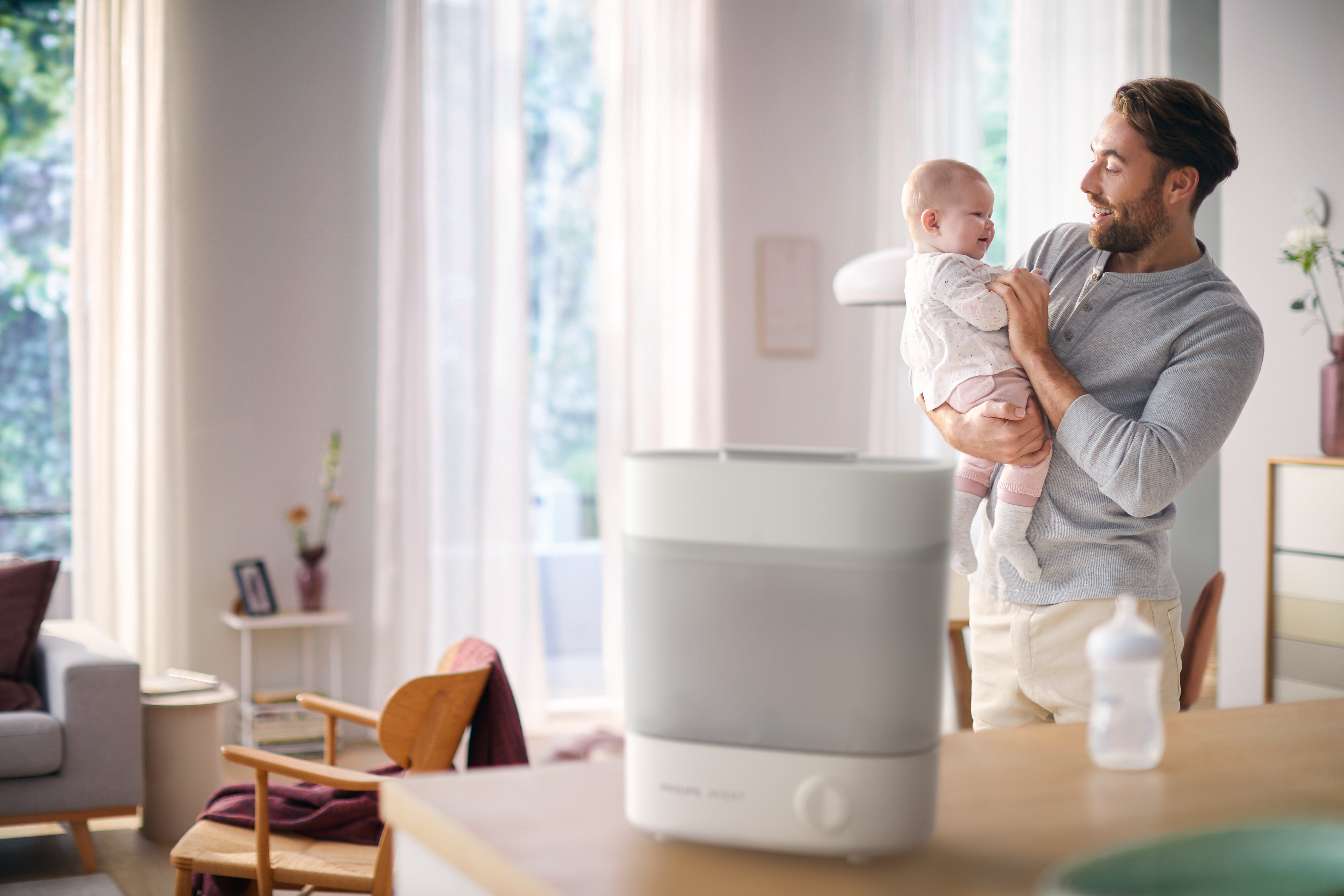 Ein Vater hält ein Baby. Ein Sterilisator und eine Flasche sind im Haus sichtbar.