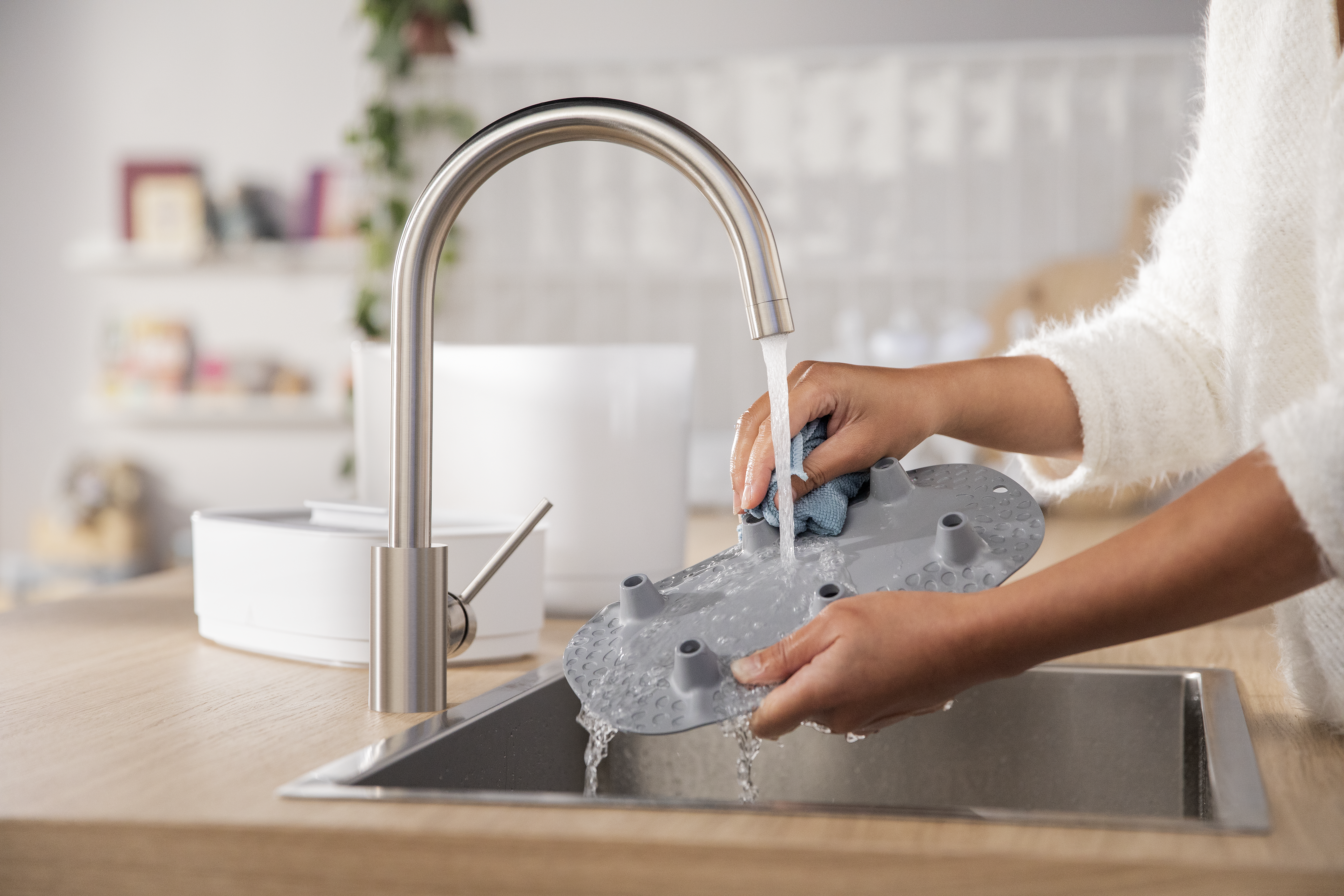 Person wäscht ein graues Babyflaschenteil in einem Waschbecken. Wasser fließt aus einer Chromarmatur. Weißer Hintergrund.