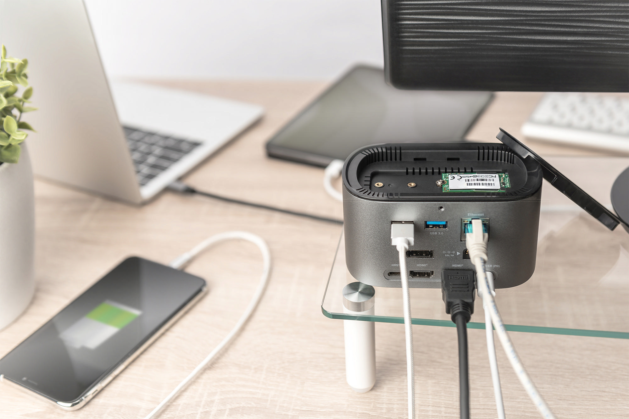 Ein ladendes Telefon, Laptop und Dock mit Kabeln auf einem Holztisch, Monitor hinten.