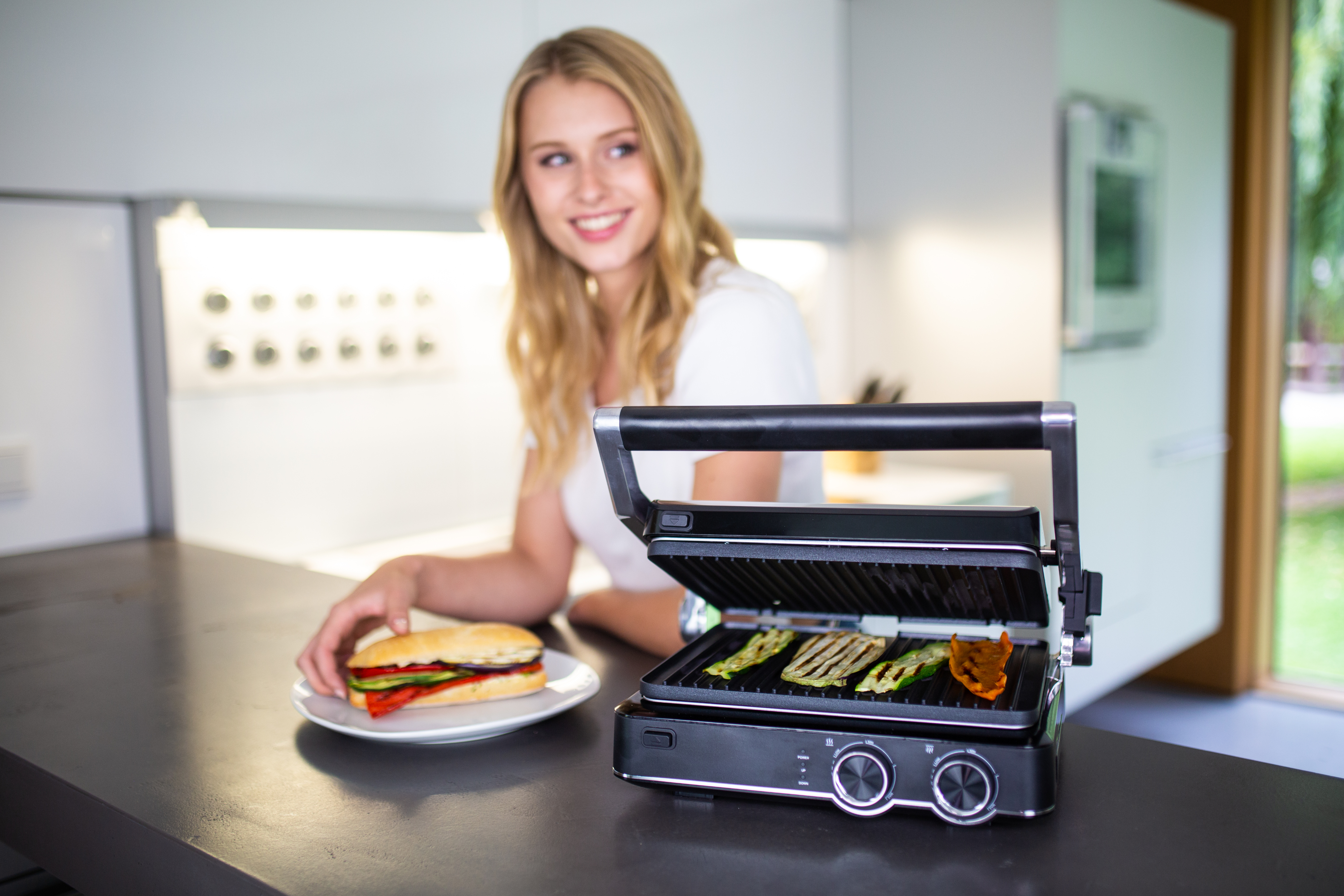 Frau lächelt an einem Grill mit Gemüse. Ein Sandwich liegt auf einem Teller neben dem Grill.