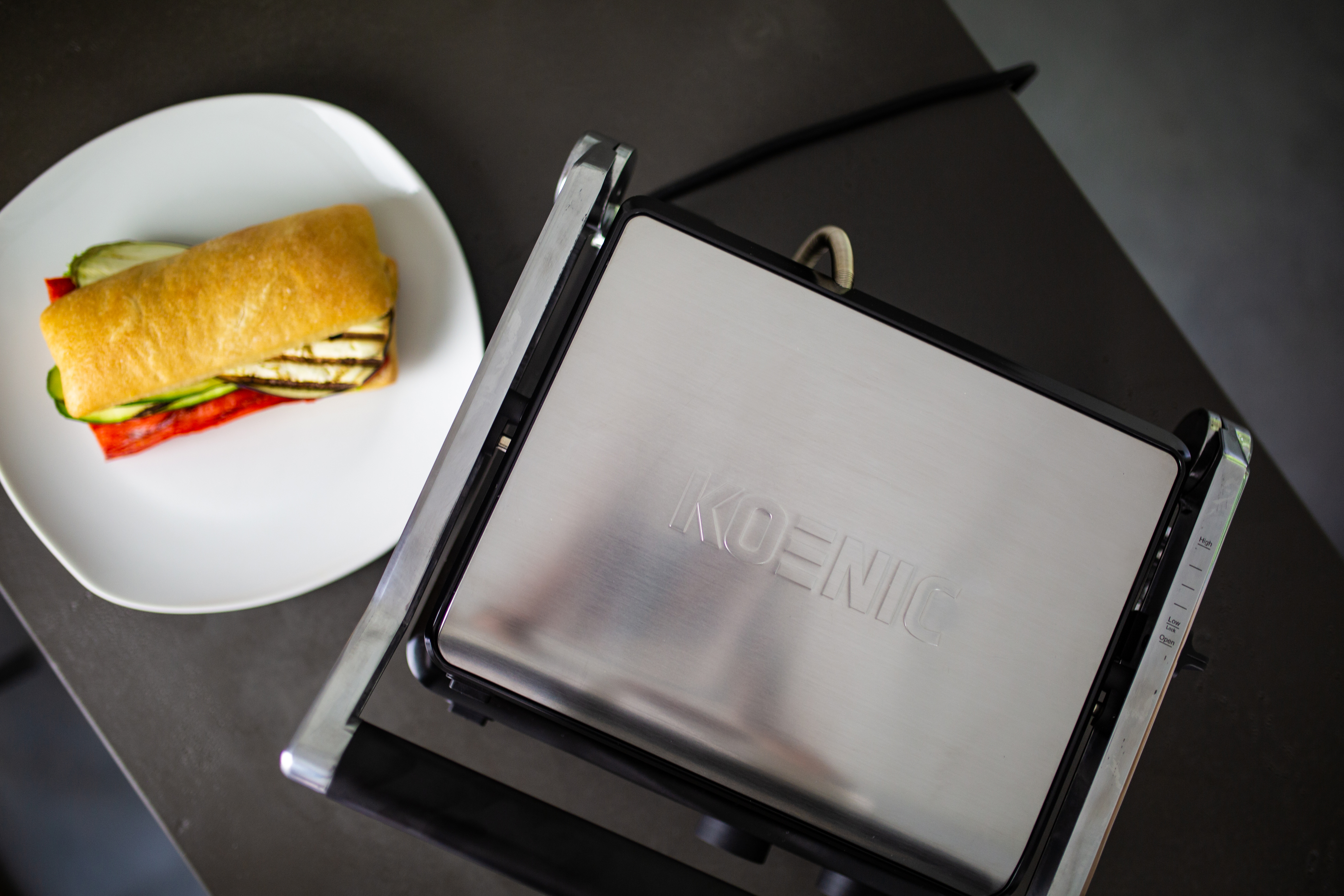 Ein Sandwich und ein Koenic-Grill. Das Sandwich hat gegrilltes Brot, Gemüse und Käse.