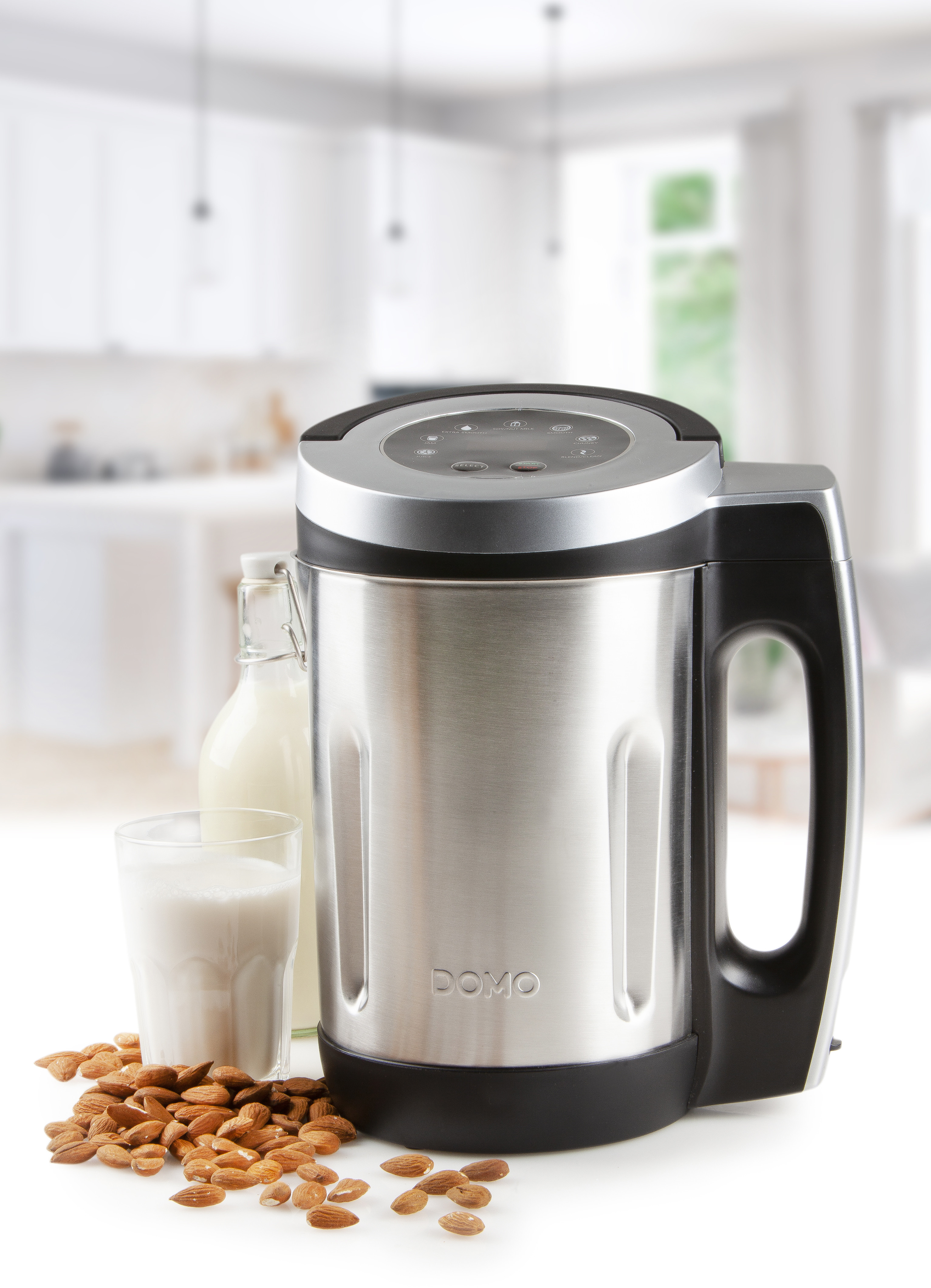 Silver blender, glass of milk, bottle of milk, and almonds sit on a white surface, kitchen background.