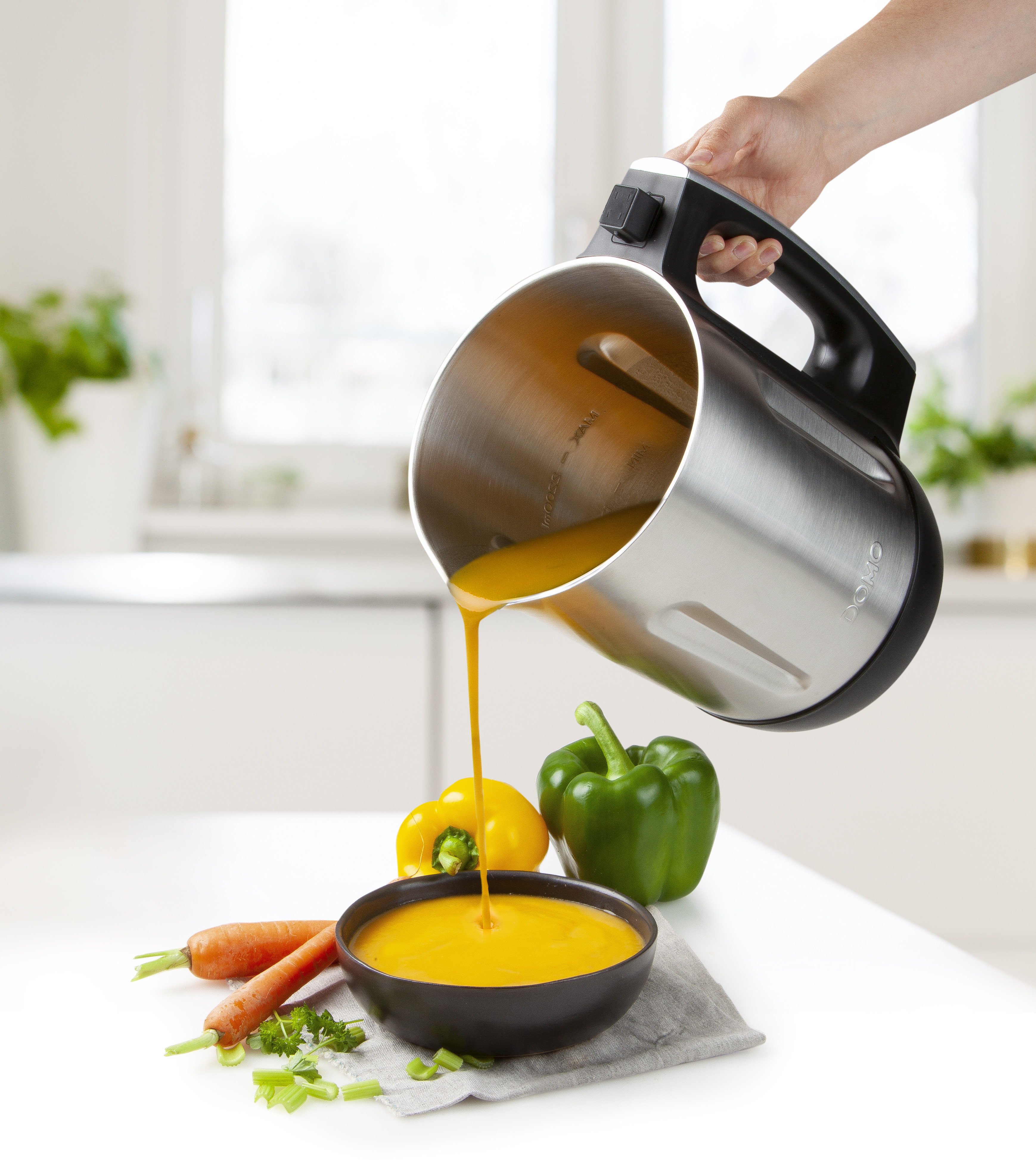 A person pouring orange soup from a silver blender into a black bowl. Vegetables are present.