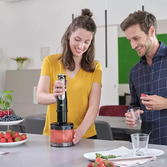 Une femme mélangeant des fraises. Un homme regarde avec un verre. Des assiettes blanches avec des fruits sur la table.