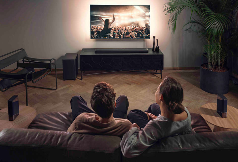 Zwei Personen auf einem Sofa schauen fern. Der Fernseher zeigt ein Konzert an der Wand. Lautsprecher an den Seiten.
