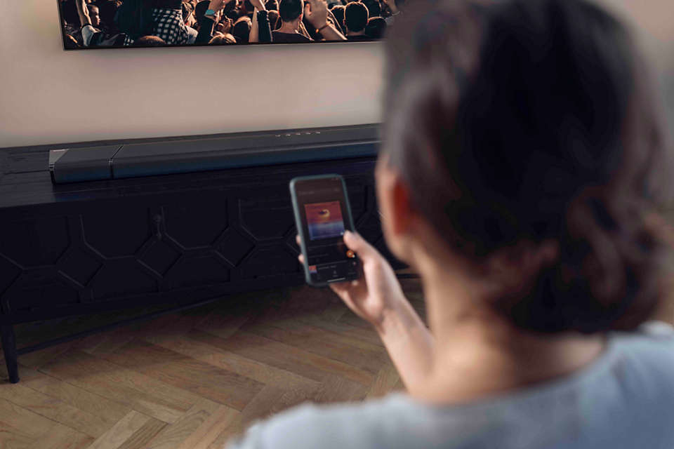 Frau hält ein Telefon vor einer Soundbar und einem Fernseher.