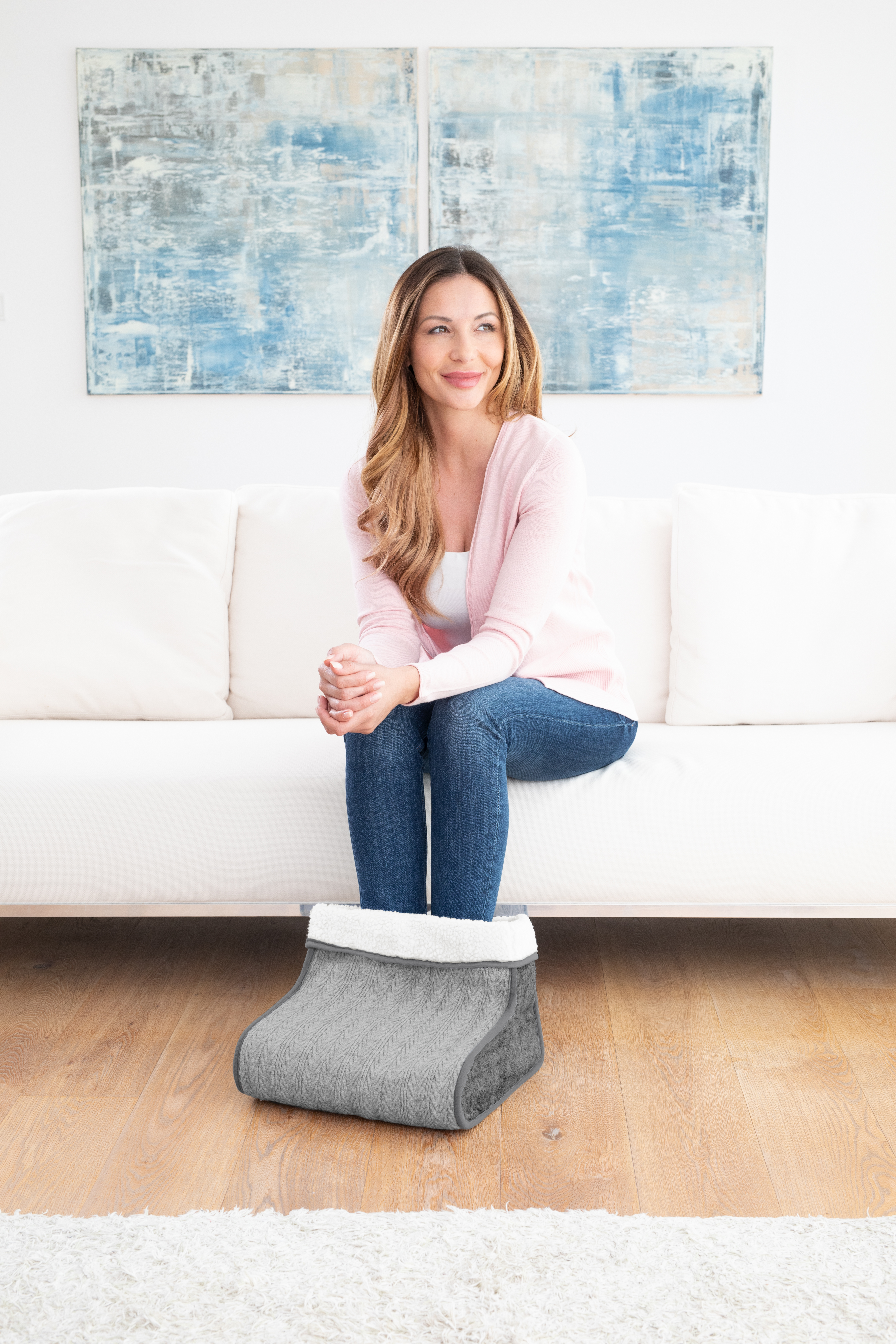 Frau sitzt auf weißem Sofa, Füße in grauem Fußwärmer. Blaue Kunstwerke im Hintergrund.