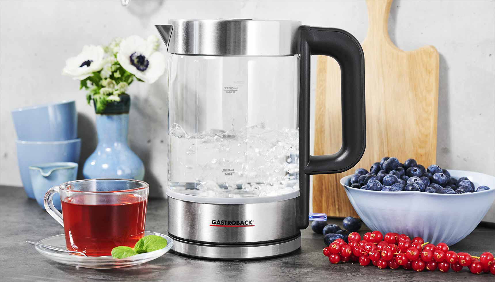Ein klarer Wasserkocher mit kochendem Wasser, Beeren, Blumen und Teetasse auf einer Küchenzeile.