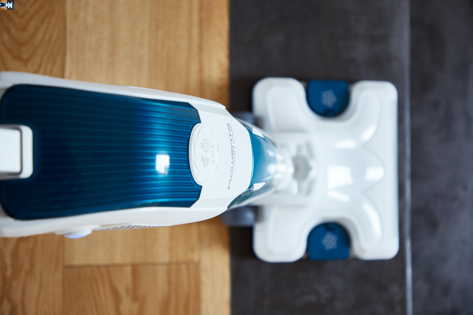 Un nettoyeur vapeur blanc et bleu sur un plancher en bois franc, avec une surface gris foncé.