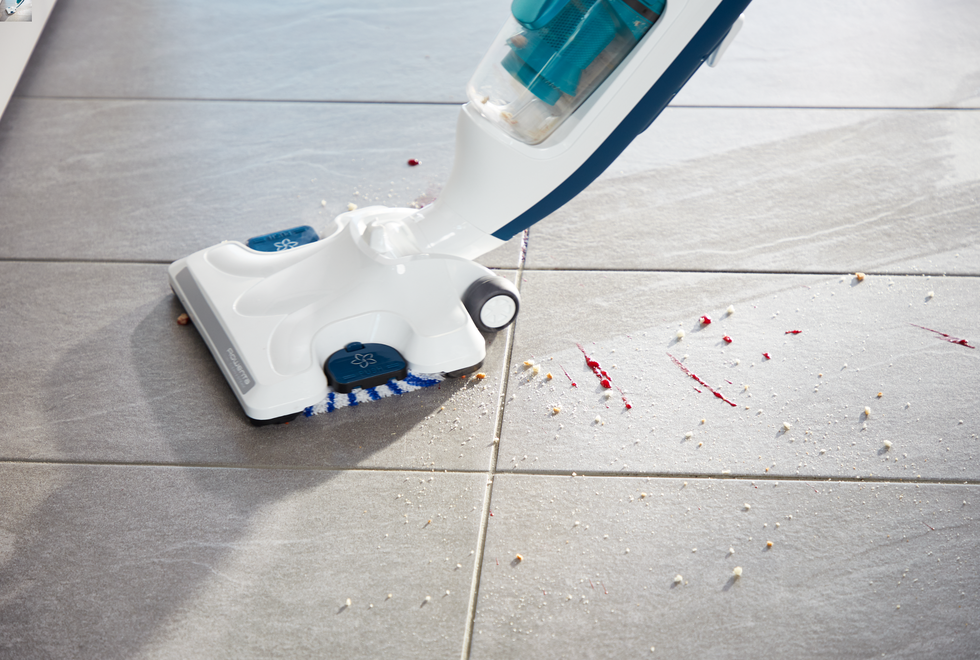 Une vadrouille à vapeur blanche et bleue nettoyant un sol en carrelage gris avec des débris et des taches rouges.
