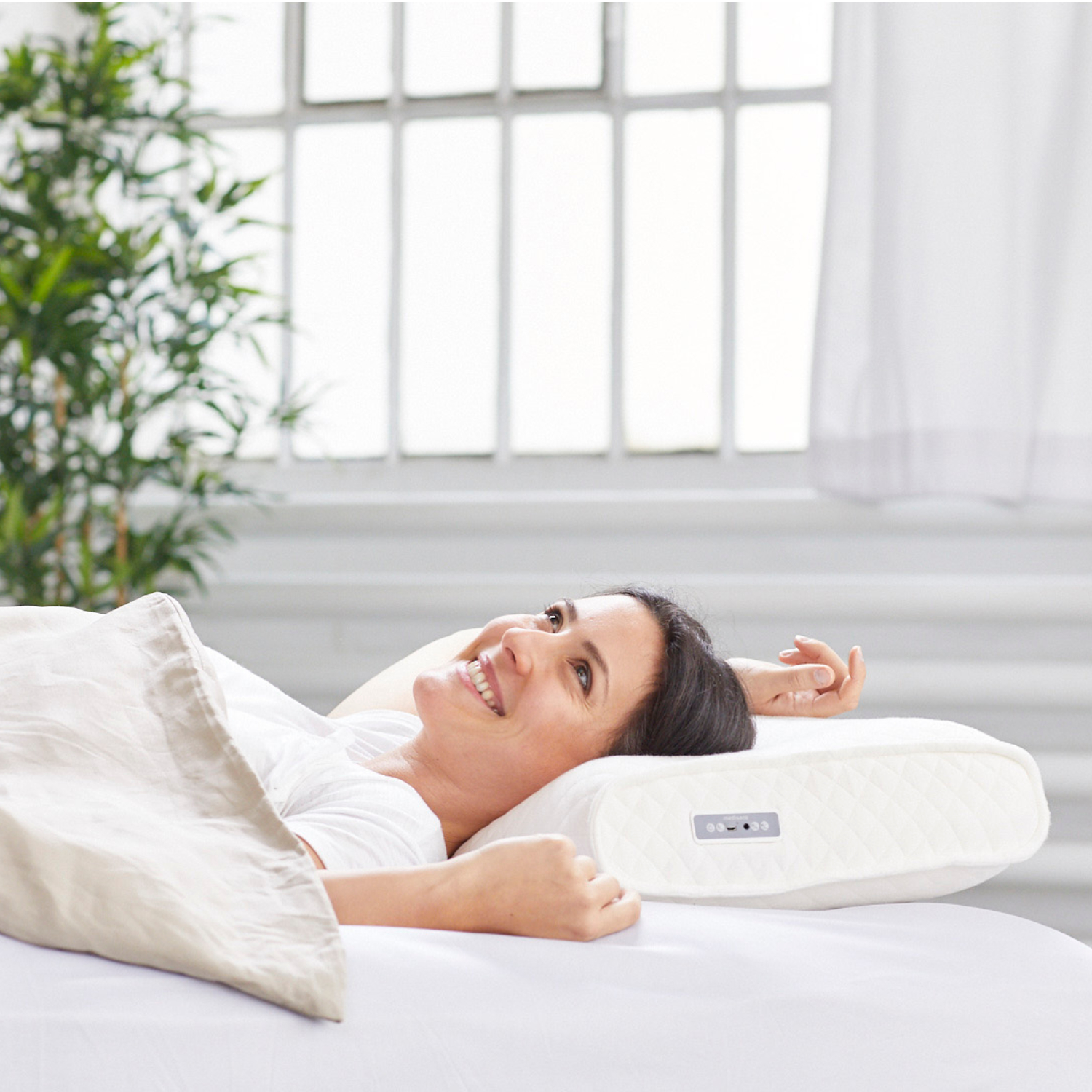 Een vrouw glimlacht terwijl ze in bed ligt op een wit kussen. Ze is bedekt met een beige deken. Een raam en planten zijn op de achtergrond.