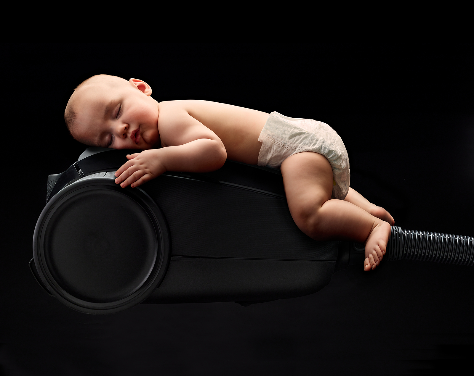 Ein Baby schläft auf einem schwarzen Staubsauger. Das Baby trägt eine Windel. Schwarzer Hintergrund.