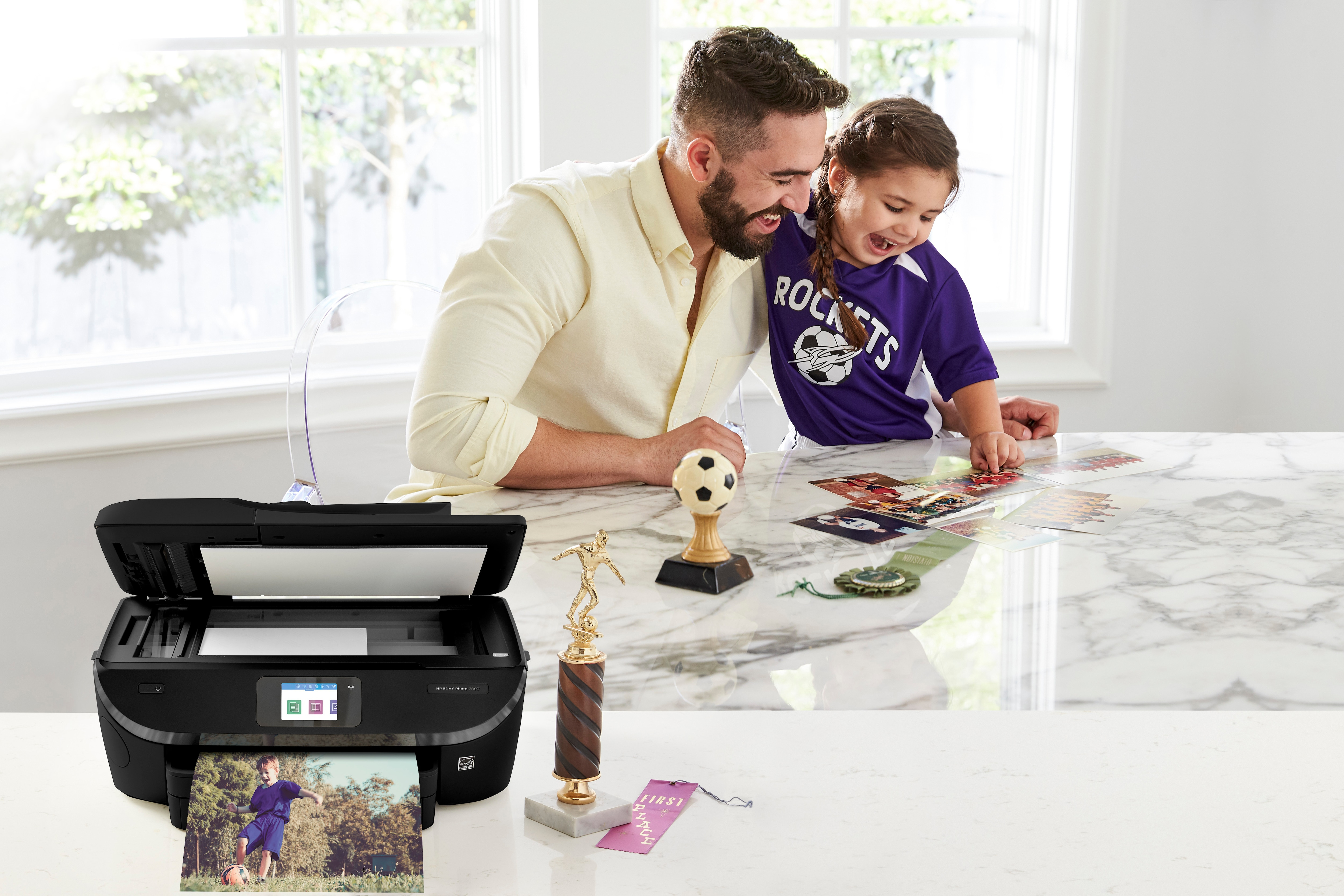 Vater und Tochter betrachten Fotos, die von einem schwarzen Drucker ausgedruckt wurden. Eine Trophäe und ein Fußball stehen auf dem Tisch.